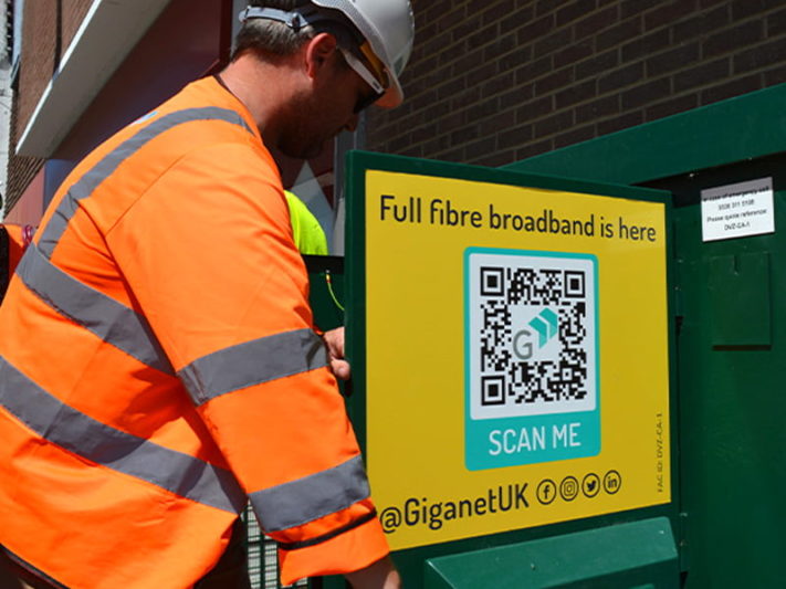 Giganet engineer installing full fibre broadband cabinet in Wimborne Giganet engineer wearing orange high-vis jacket installing green full fibre broadband cabinet in Wimborne